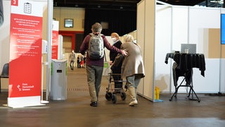 Ein Mann geleitet eine ältere Frau mitRollator ins Bremer Impfzentrum