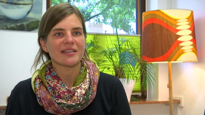 Eine junge Frau schaut in die Kamera, im Hintergrund stehen Pflanzen und eine Lampe.