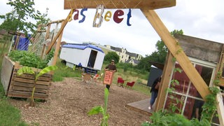 Hinter einem Holzbogen mit der Aufschrift "Das Beet" stehen Container und Menschen.