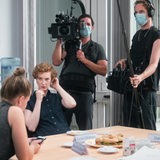 Zwei Frauen und ein Mann sitzen an einem Tisch, dahinter stehen zwei Männer mit Kamera und Mikrofon.
