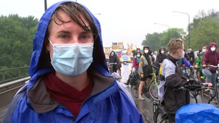 Eine Frau mit Regenjacke, nassen Haaren unter einer Kapuze und Mundschutzmaske. Dahinter viele Radfahrer.