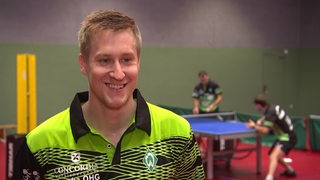 Mattias Falck lächelt beim Interview mit dem Sportblitz, im Hintergrund trainieren zwei Werder-Spieler.