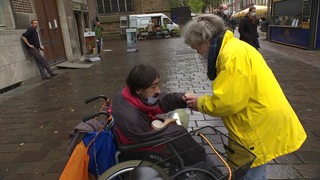 Frau mit gelber Regenjacke überreicht einem im Rollstuhl sitzenden Obdachlosen einen Kaffe.