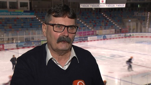 Pinguins-Manager Alfred Prey steht in der Eishalle beim Interview, während die Mannschaft hinter ihm auf dem Eis trainiert.