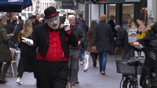 Ein geschminkter Mann läuft eine Straße entlang und hält sich einen Handfeger wie ein Telefon ans Ohr.