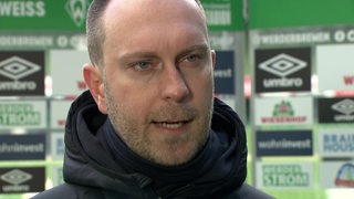 Werder-Trainer Ole Werner steht nach dem Spiel vor einer Werbewand beim Interview.