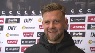 Werder-Stürmer Niclas Füllkrug steht mit breitem Grinsen vor einer Werbewand beim Interview.