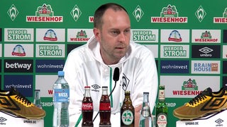 Werder-Coach Ole Werner im weißen Kapuzenshirt vor einer Werbewand beim Interview.