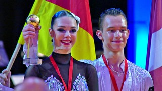 Luna Albanese und Dimitri Kalistov stehen strahlend auf dem Siegerpodest mit ihrem Pokal nach dem Gewinn der Junioren-WM.
