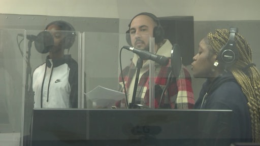 Drei Personne stehen im Tonstudio vor ihren Mikros und singen einen Song ein 