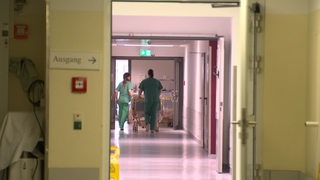 Zwei Krankenhausmitarbeiterinnen schieben ein Bett über einen Klinikflur.
