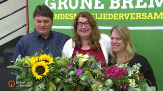 Dietmar Strehl, Anja Stahmann und Maike Schaefer zusammen auf dem Landesparteitag der Grünen. In ihren Händen haben sie alle einen Blumenstrauß.