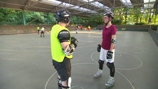 Reporterin Maike Albrecht mit Inline-Skates in einer Sporthalle auf einem Hockey-Feld, die sich mit einem Hockey-Spieler unterhält. Im Hintergrund weitere Hockey-Spieler im Training