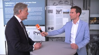 Reporter János Kereszti steht rechts im Bild, er interviewt den Historiker und Publizist Norbert Frei und hält ihm ein Mikrofon hin.