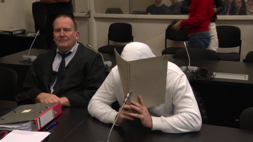 Der Angeklagte aus Bremerhaven sitzt im Oberlandesgericht in Hamburg und hält eine Mappe vor sein Gesicht. 