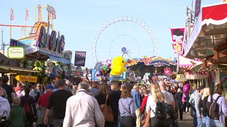 Viele Menschen laufen über das Gelände des Ischa Freimarkts, mit vielen Fahrgeschäften und einem Riesenrad im Hintergrund.