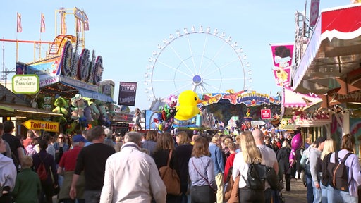 Viele Menschen laufen über das Gelände des Ischa Freimarkts, mit vielen Fahrgeschäften und einem Riesenrad im Hintergrund.