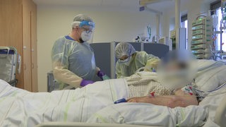 Zwei Menschen stehen neben einem Krankenhausbett und kümmern sich um eine Covid-19-Patientin.