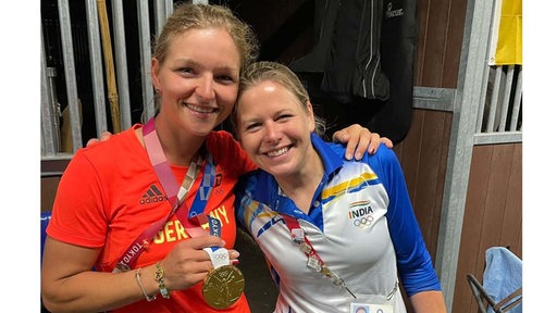 Zwei blonde Frauen lachen in die Kamera. Die eine zeigt ihre goldene Olympia-Medaille
