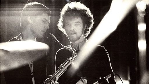 Schwarzweiß-Foto zweier Männer, einer mit Saxophon, in Scheinwerferlicht