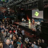 Publikum in vollem Auditorium blickt herunter auf Bühne mit Musikern