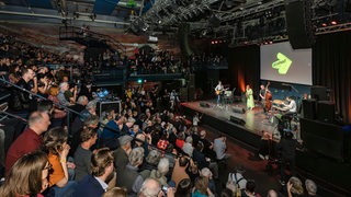 Publikum in vollem Auditorium blickt herunter auf Bühne mit Musikern