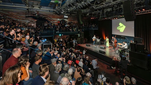 Publikum in vollem Auditorium blickt herunter auf Bühne mit Musikern