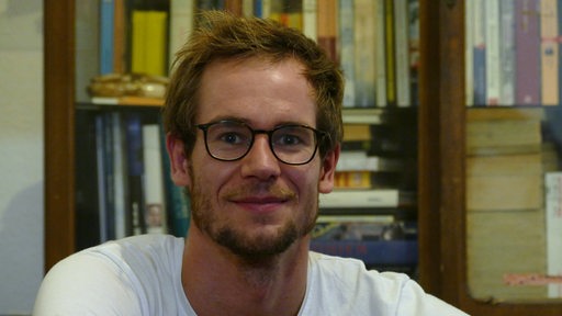 Ein Mann mit Brille lächelt in die Kamera. Er sitzt vor einem Schrank gefüllt mit Büchern.