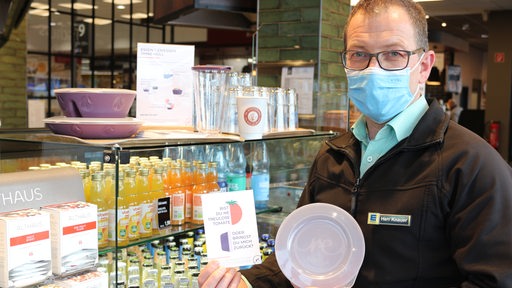 Jens Knauer steht mit einer Plastikdose und einem Flyer vor einer Getränkevitrine
