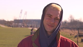 Ein junger Mann mit einer Kapuze auf dem Kopf lächelt in die Kamera und blinzelt in der Sonne.