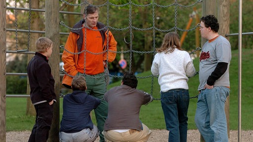 Ein Sozialpädagoge steht mit Jugendlichen und Kindern auf einem Spielplatz