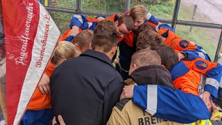 Eine Gruppe Kinder und Erwachsene in Uniform stehen mit verschlungenen Armen im Kreis.