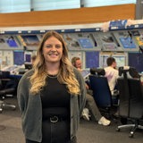 Julia Schult steht vor der Brücke im Radarkontrollzentrum und lächelt in die Kamera. Ihre Arme hat sie hinter dem Rücken. Im Hintergrund sieht man ihre Kolleginnen und Kollegen unscharf.