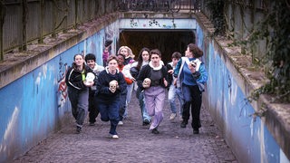 Gruppe Jugendlicher, einige mit Schädel in der Hand, sprinten aus Fußgängertunnel Richtung Kamera