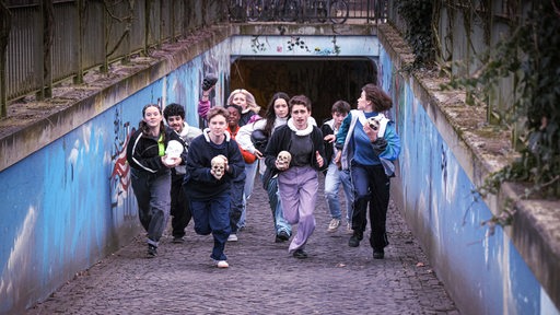 Gruppe Jugendlicher, einige mit Schädel in der Hand, sprinten aus Fußgängertunnel Richtung Kamera