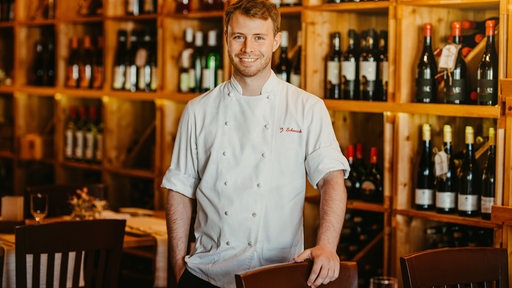 Junger Mann mit Schürze, offenbar Koch, posiert vor Weinregal in einem Restaurant