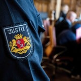 Ein Justizbeamter steht im Gerichtssaal, während der Angeklagte vor Prozessbeginn sein Gesicht hinter einer Mappe verbirgt. 