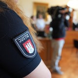 Eine Justizbeamtin steht im Sitzungssaal, auf ihrem Arm steht "Justiz".