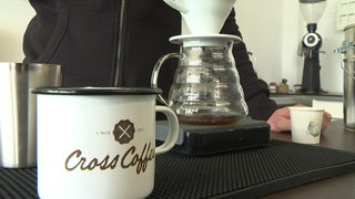 Eine Kaffeetasse mit der Aufschrift " Cross Coffee". Dahinter bereitet jemand einen Filterkaffee zu.