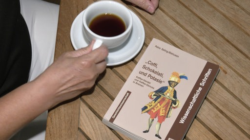 Eine Hand greift nach einer Kaffeetasse, die auf einem Tisch neben einem Buch steht.