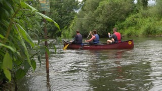 Drei Personen sitzen in einem Kanu auf einem Gewässer. Im Hintergrund sind viele grüne Bäume.