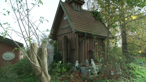 Ein Mann in Mönchskostüm steht vor dem  Nachbau einer hölzernen Kapelle