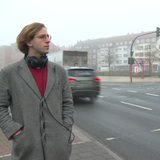 Karl Geibert steht mit Kopfhörern an einer stark befahrenen Hauptstraße.