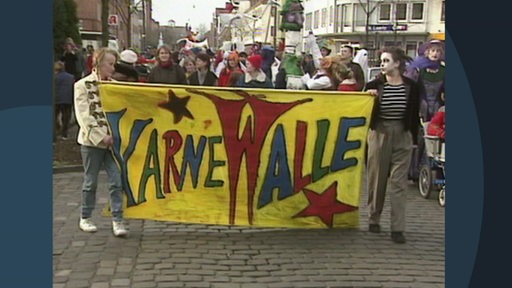 Einige Karnevalsjecken tragen ein Banner mit dem Schriftzug Karne-walle.