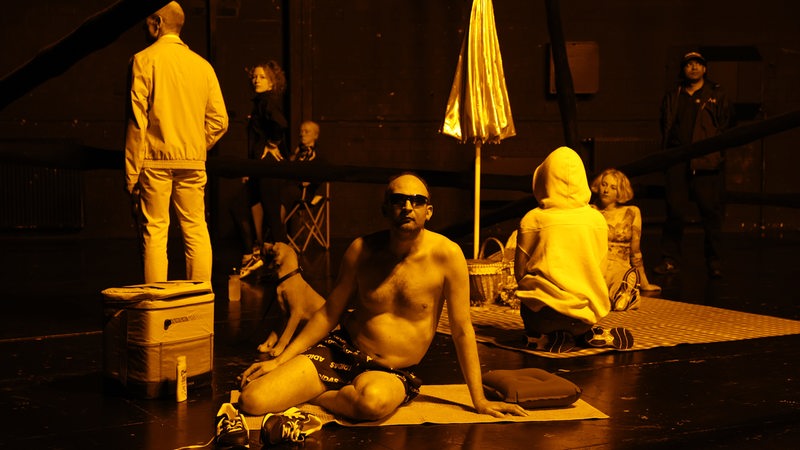 Auf einer Bühne stehen und sitzen Menschen in die Kamera schaut ein Mann in Badehose mit Sonnenbrille