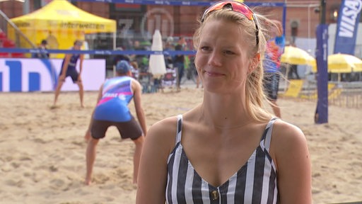 Die Beachvolleyballspielerin Kim Behrens bei einem Interview und im Hintergrund ein Beachvolleyballfeld mit Spielern.