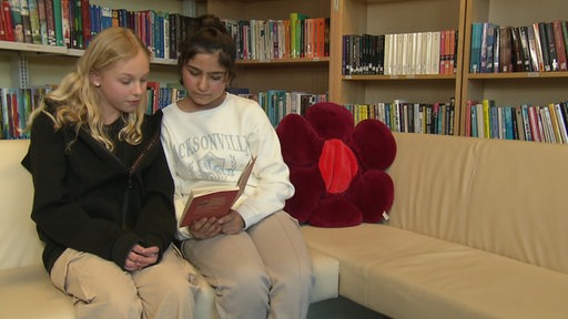 Zwei junge Mädchen sitzen nebeneinander auf einem Sofa und lesen aus einem Buch. Im Hintergrund Regale, voll mit Büchern.