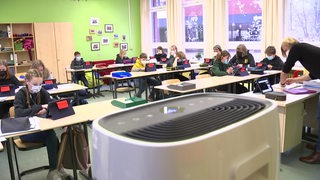 Schüler und Schülerinnen sitzen mit Masken und Tablets im Klassenraum, im Vordergrund ein Luftfilter. 