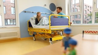 Ein Mädchen sitzt im Krankenhausbett, ein Arzt sitzt neben ihr