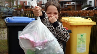 Ein Mädchen bringt den Müll in die Mülltonne und hält sich dabei lachend die Nase zu.